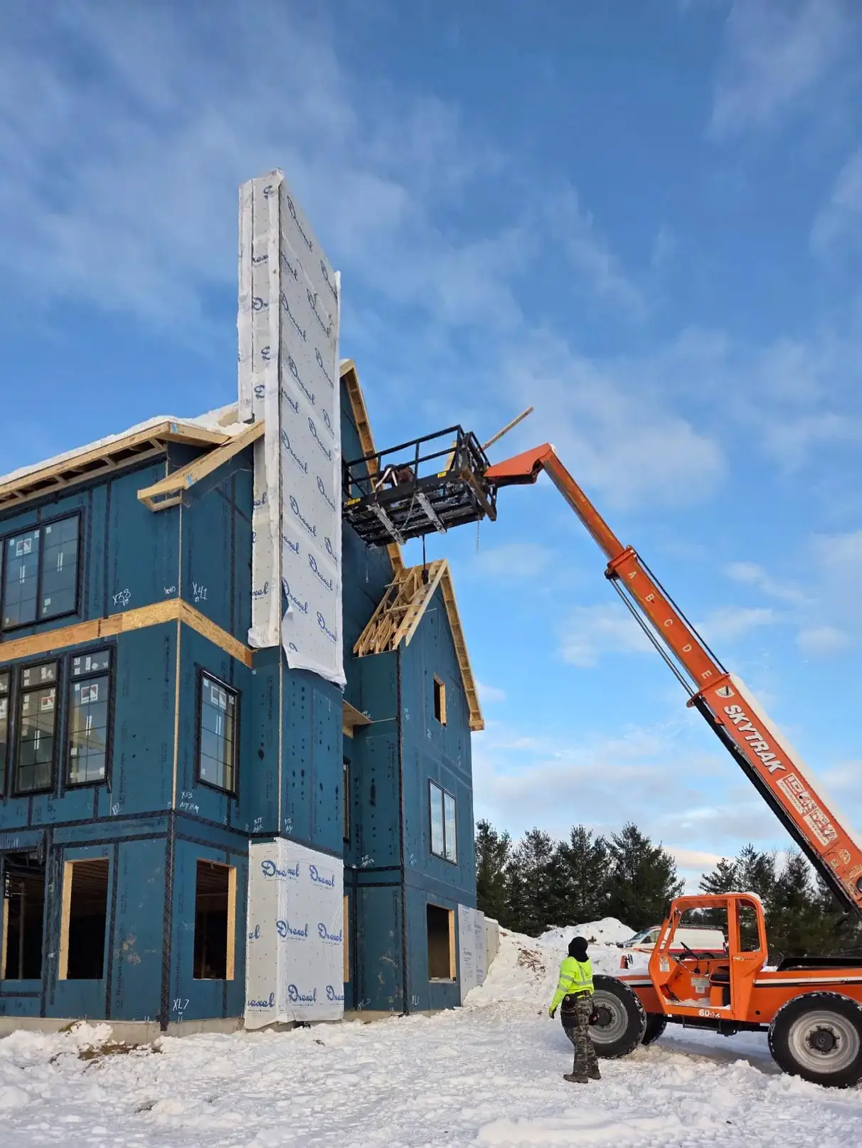 Two story new home with a carpenter on a lift on the left hand side of the home. Snow on the ground around newly framed home.